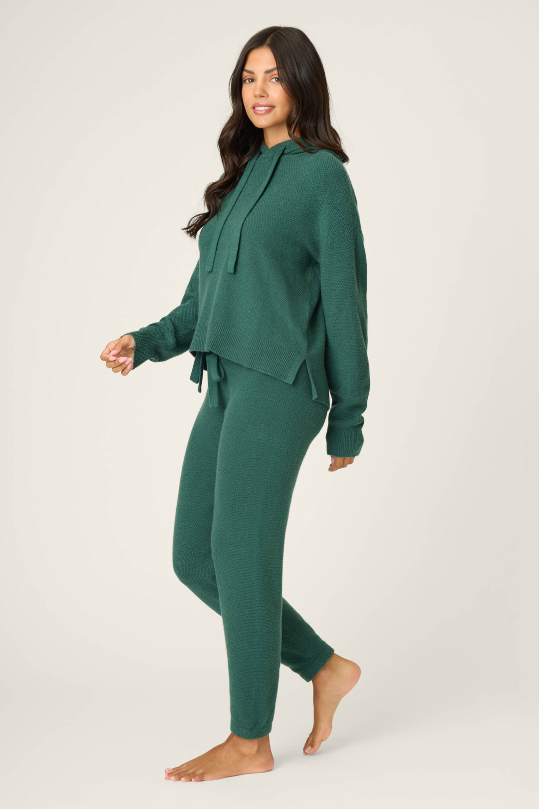 A woman with long dark hair stands barefoot on a light background, smiling and wearing the P.J. Salvage SOLID PJ SET SWEATER WEATHER—matching dark green knit hoodie sweater and fitted pants. She is turned slightly to the side.