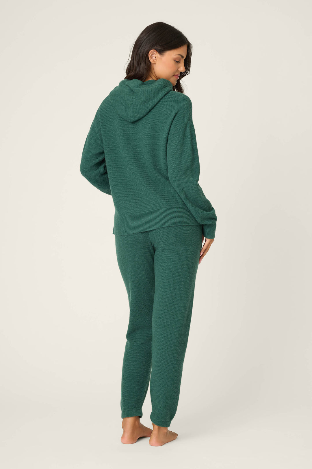 A person stands barefoot on a plain light background, facing away, wearing the P.J. Salvage SOLID PJ SET SWEATER WEATHER in dark green. Their head is turned slightly to the side and their hair is down.