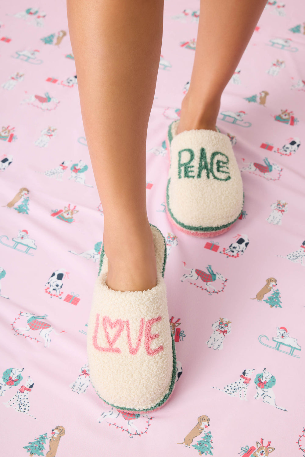 A person wears P.J. Salvage LOVE PEACE SLIPPER PEACE & PAWLIDAYS on a pink dog-patterned sheet; one slipper reads LOVE in pink, the other PEACE in green.