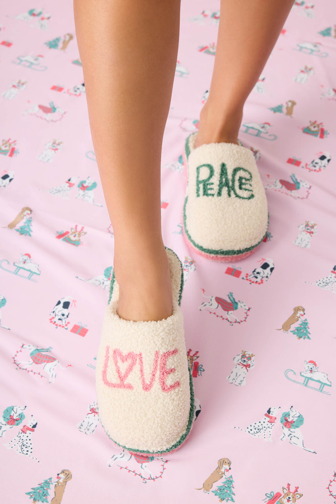 A person wears P.J. Salvage LOVE PEACE SLIPPER PEACE & PAWLIDAYS on a pink dog-patterned sheet; one slipper reads LOVE in pink, the other PEACE in green.