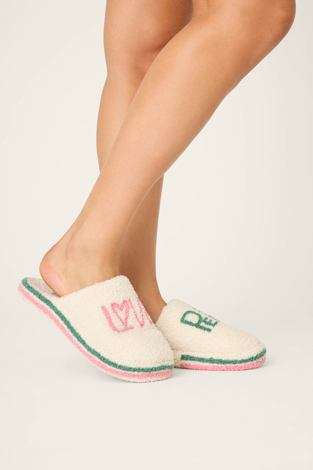A person wears P.J. Salvage LOVE PEACE SLIPPER PEACE & PAWLIDAYS: fuzzy cream slippers with green and pink trim; the left shows "LOVE" in pink, the right "PEACE" in green. The background is plain and light-colored.