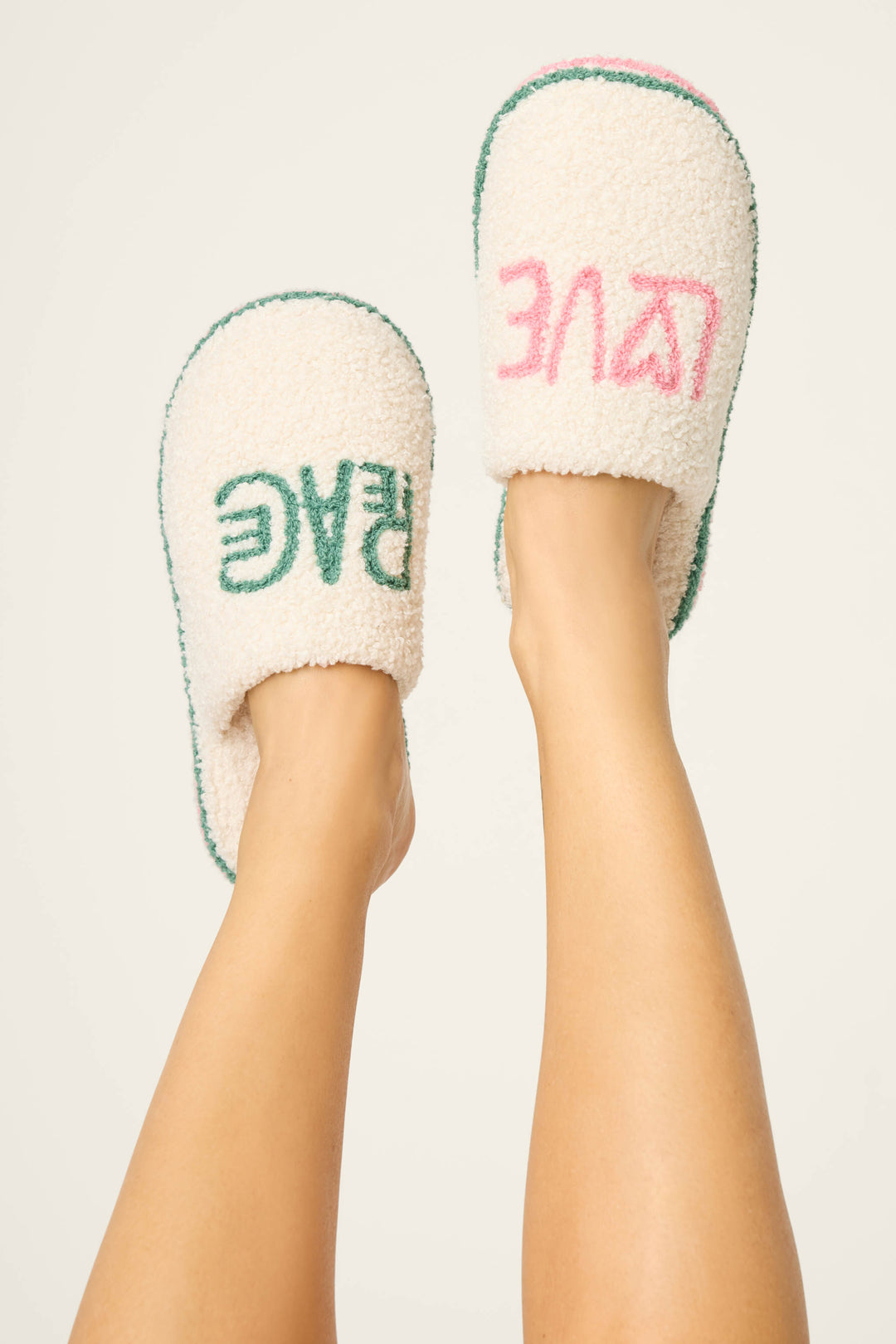 A pair of legs raised upward wear P.J. Salvage LOVE PEACE SLIPPER PEACE & PAWLIDAYS fuzzy slippers—one reads "PEACE" in green, the other "LOVE" in pink (text reversed)—against a plain, light background.