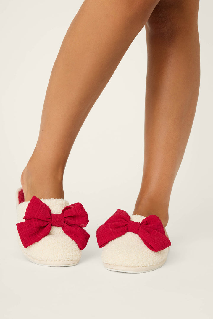 A person stands on a light surface, showing only their lower legs and feet while wearing P.J. Salvage BOW SLIPPER FELINE FESTIVE—white fuzzy slippers adorned with large red bows.