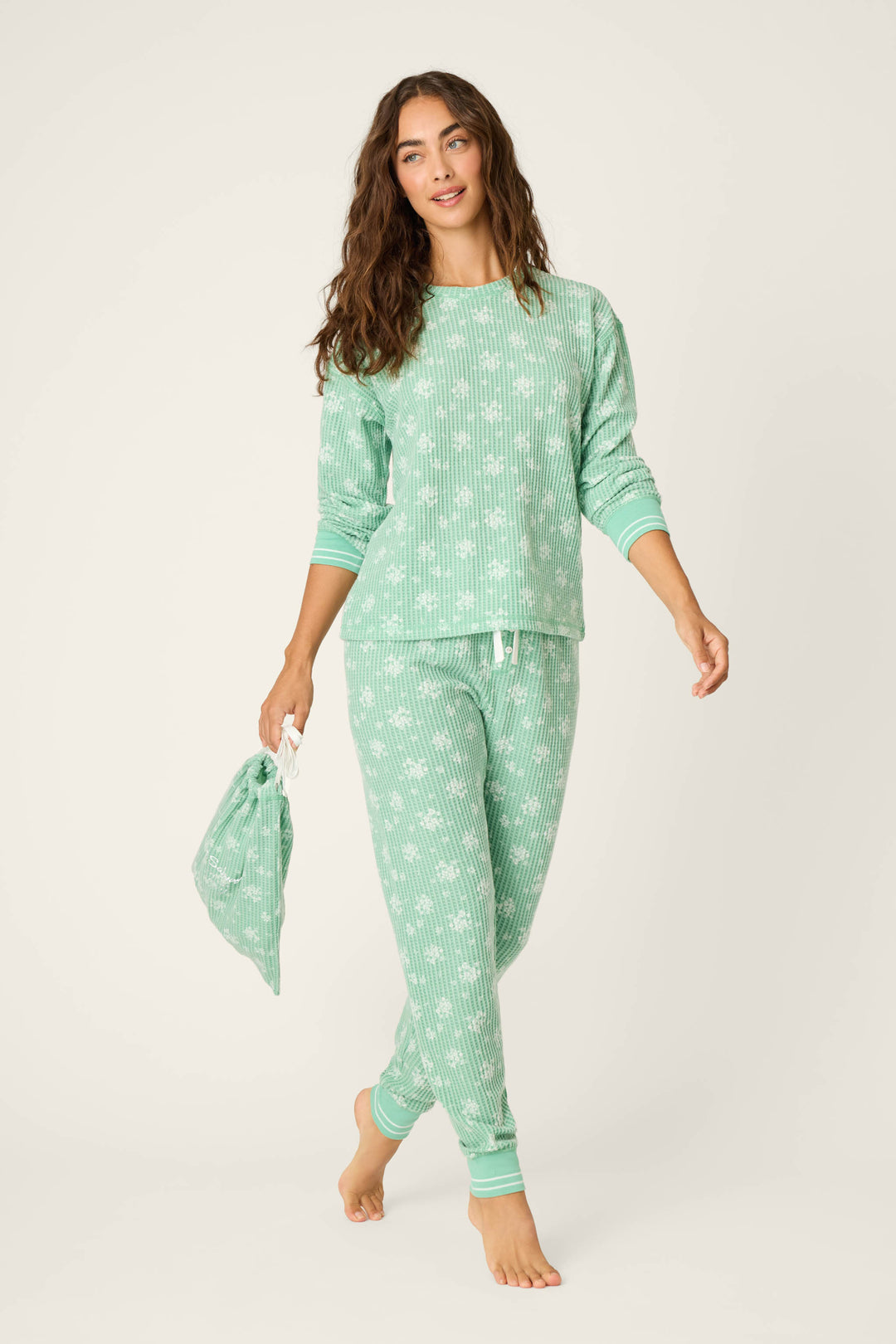 A woman with long brown hair wears the P.J. Salvage DTC Exclusive - SNOWFLAKE PJ SET 3 PIECE GIFT SET in light green with a white pattern, barefoot and holding a matching drawstring bag against a plain light background.