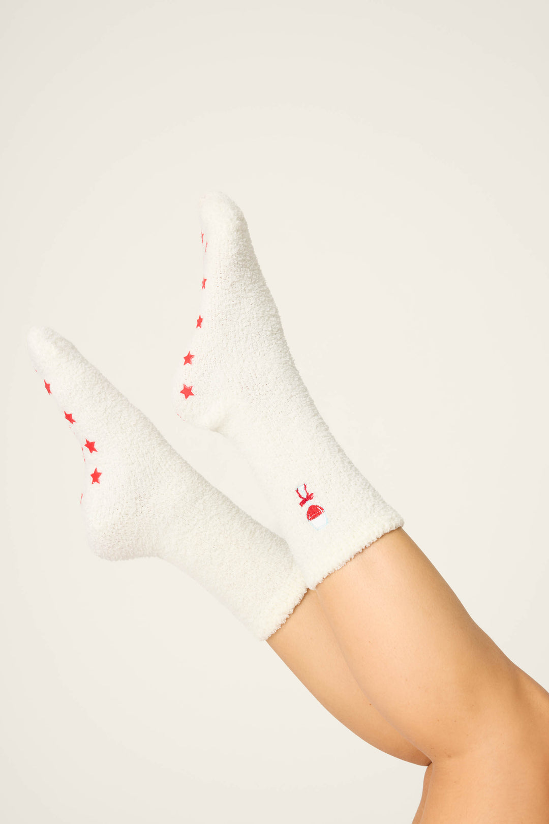 A person’s legs are raised, wearing P.J. Salvage DRINK SOCKS LET'S GET TOASTY—white fuzzy socks with red stars on the soles and a small red and white reindeer face on the sides—against a plain, light background.
