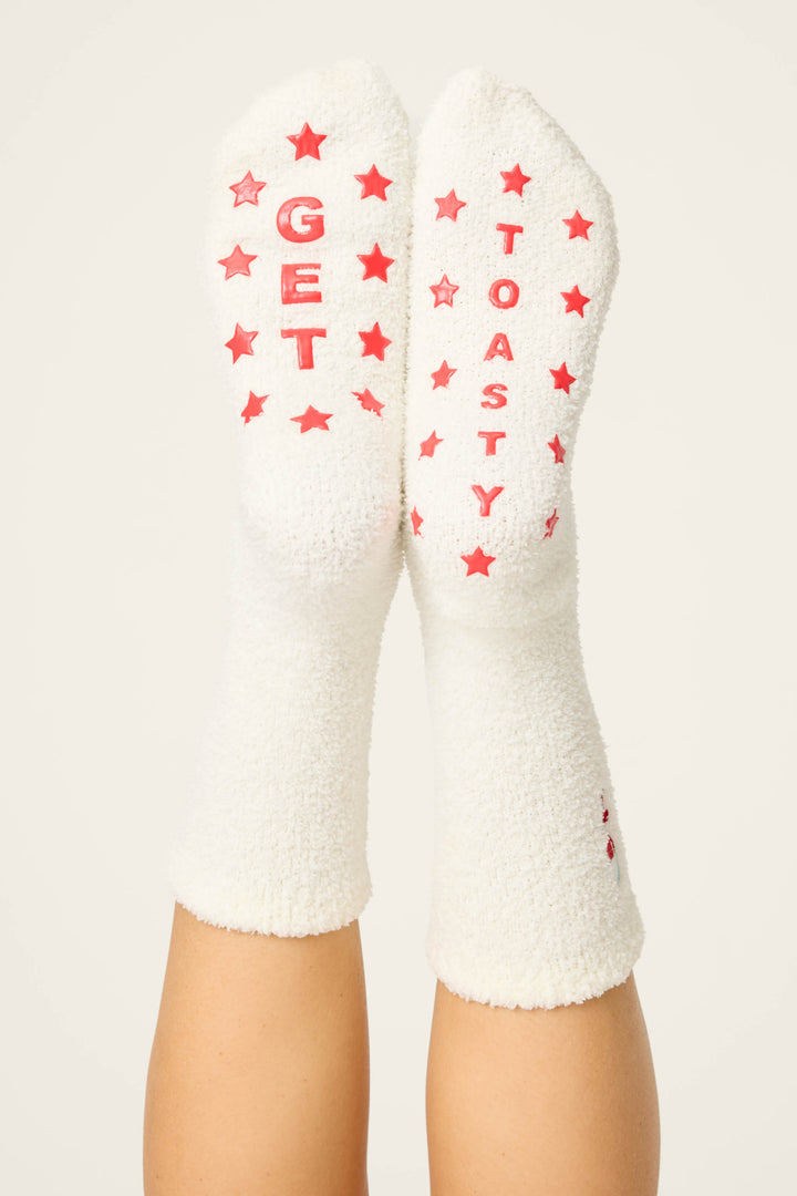 A pair of feet wear P.J. Salvage DRINK SOCKS LET'S GET TOASTY—fuzzy white socks with red stars and "GET TOASTY" in red on the soles—against a plain, off-white background.
