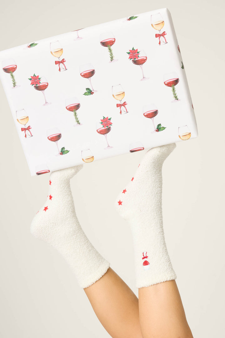 A person wears P.J. Salvage’s DRINK SOCKS LET'S GET TOASTY (white socks with red stars) and holds a gift box wrapped in paper featuring wine glasses, holly, and poinsettias against a plain, light-colored background.