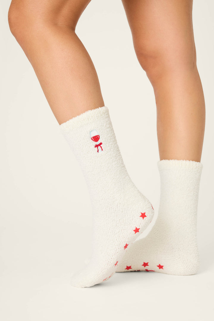 A person wears P.J. Salvage DRINK SOCKS LET'S GET TOASTY—white, fuzzy socks with red stars on the soles and a small red balloon embroidered near the top—standing against a plain light background.