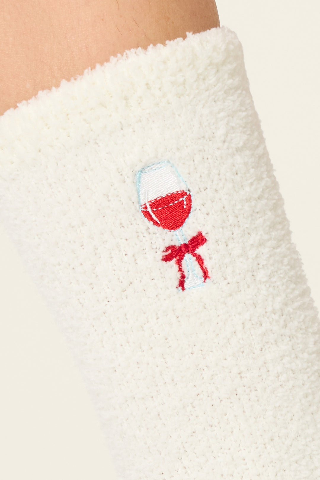 A close-up of P.J. Salvage's Let's Get Toast Wine Print Crew Socks with Grippers, featuring white textured fabric and an embroidered red wine glass with a small red bow below it.