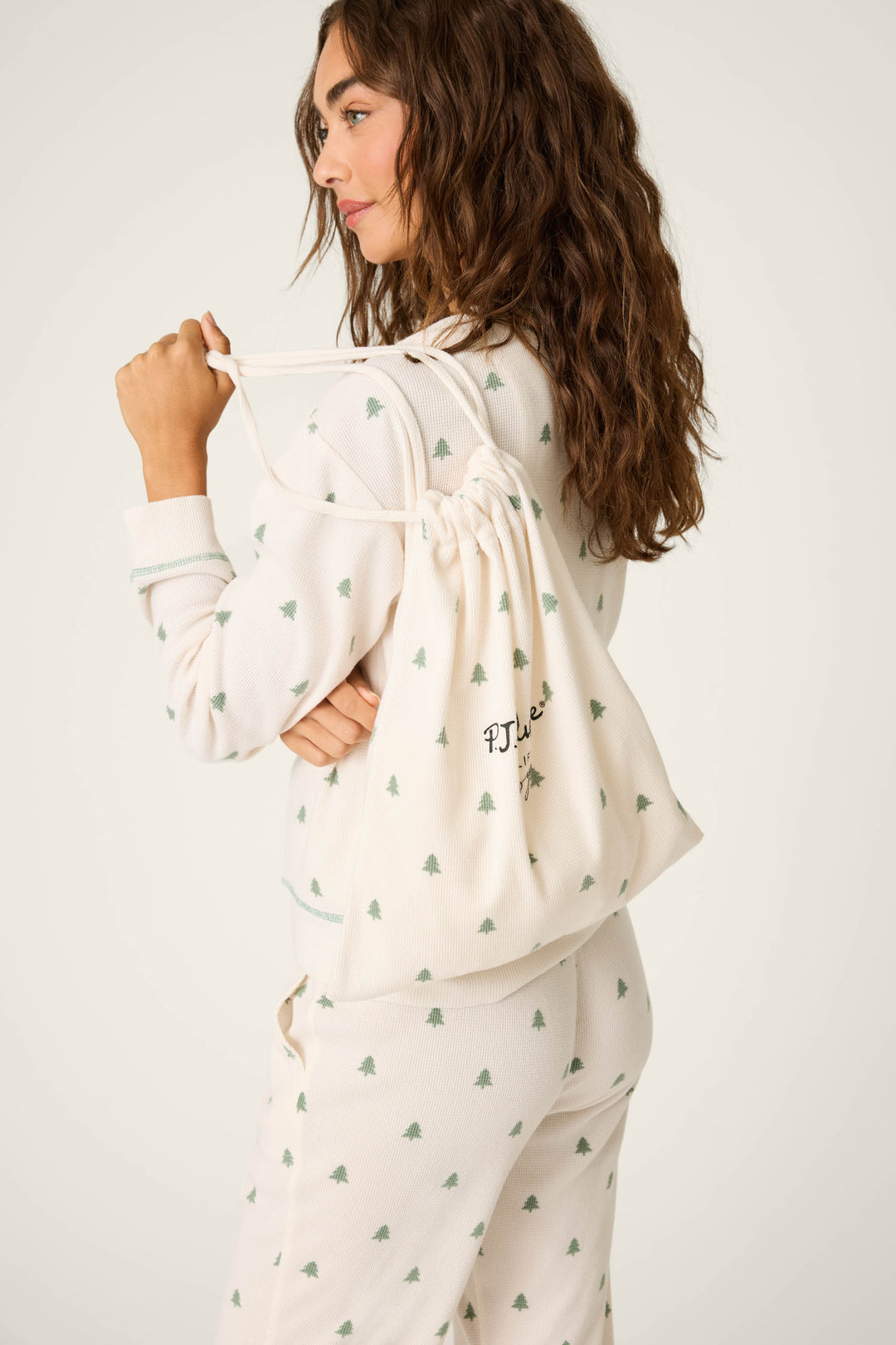 A woman with long wavy hair wears the P.J. Salvage DTC Exclusive - Plaid PJ Set 4 Piece Gift Set in white, featuring a small green tree print and carries a matching drawstring bag with P.J. Salvage embroidery over her shoulder.