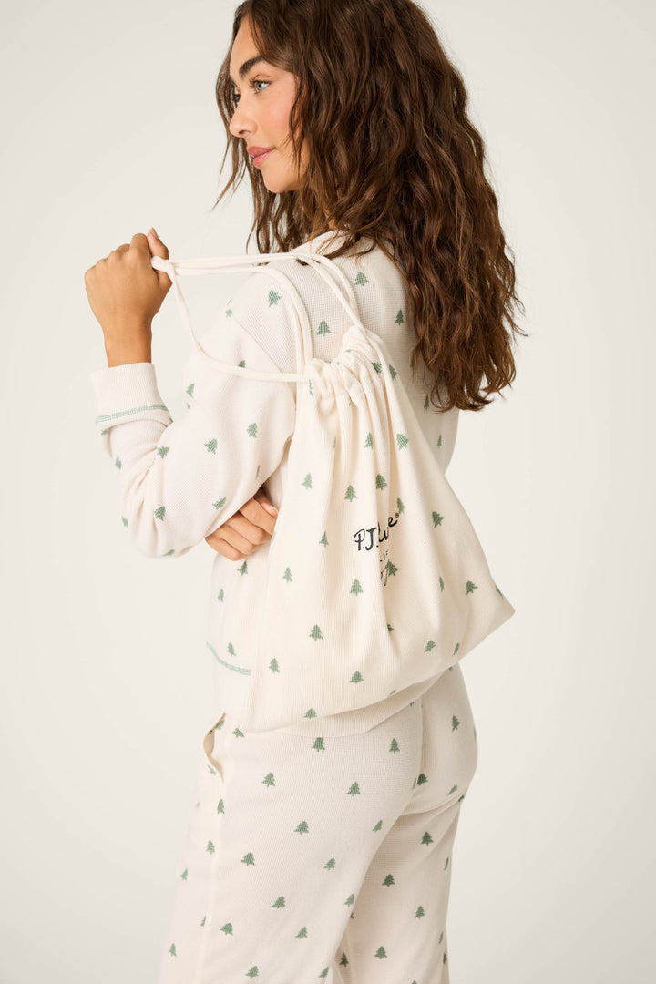 A woman with long wavy hair wears the P.J. Salvage DTC Exclusive - Plaid PJ Set 4 Piece Gift Set in white, featuring a small green tree print and carries a matching drawstring bag with P.J. Salvage embroidery over her shoulder.