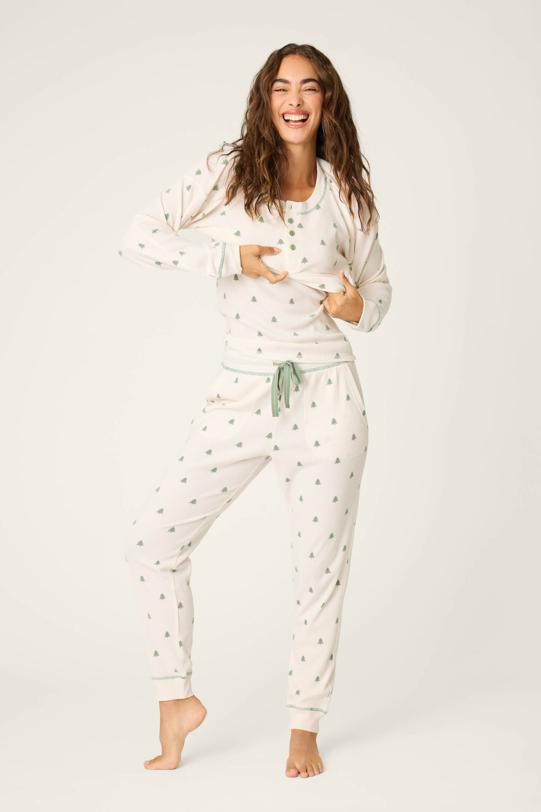 A woman stands barefoot on one leg, smiling and holding up her long-sleeve pajama top. She wears the P.J. Salvage DTC Exclusive - PLAID PJ SET 4 PIECE GIFT SET against a plain white background.