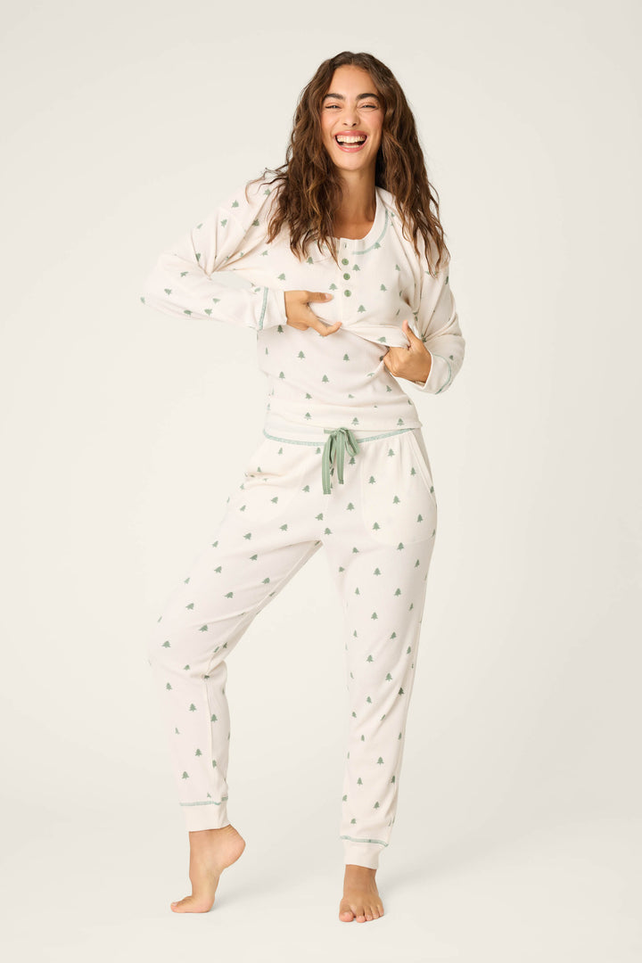 A woman stands barefoot on one leg, smiling and holding up her long-sleeve pajama top. She wears the P.J. Salvage DTC Exclusive - PLAID PJ SET 4 PIECE GIFT SET against a plain white background.