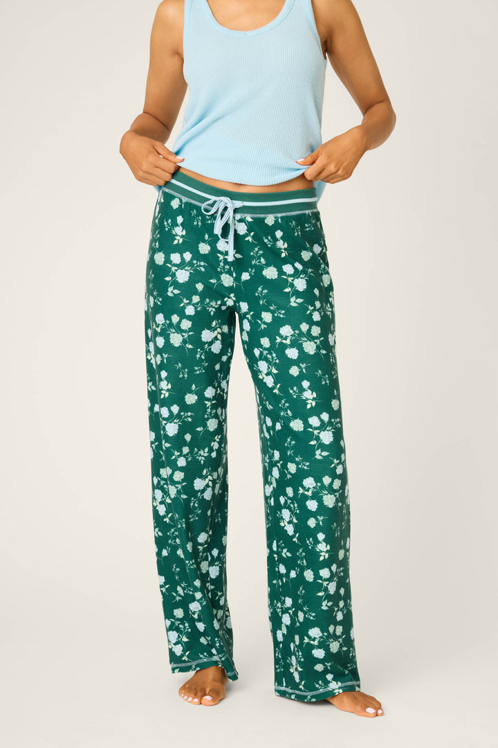 A person stands barefoot against a plain background, wearing P.J. Salvage Winter Wonderland Floral Thermal Velour Open Leg Pants with a white wintergreen floral pattern and a light blue sleeveless top, holding up the hem slightly.
