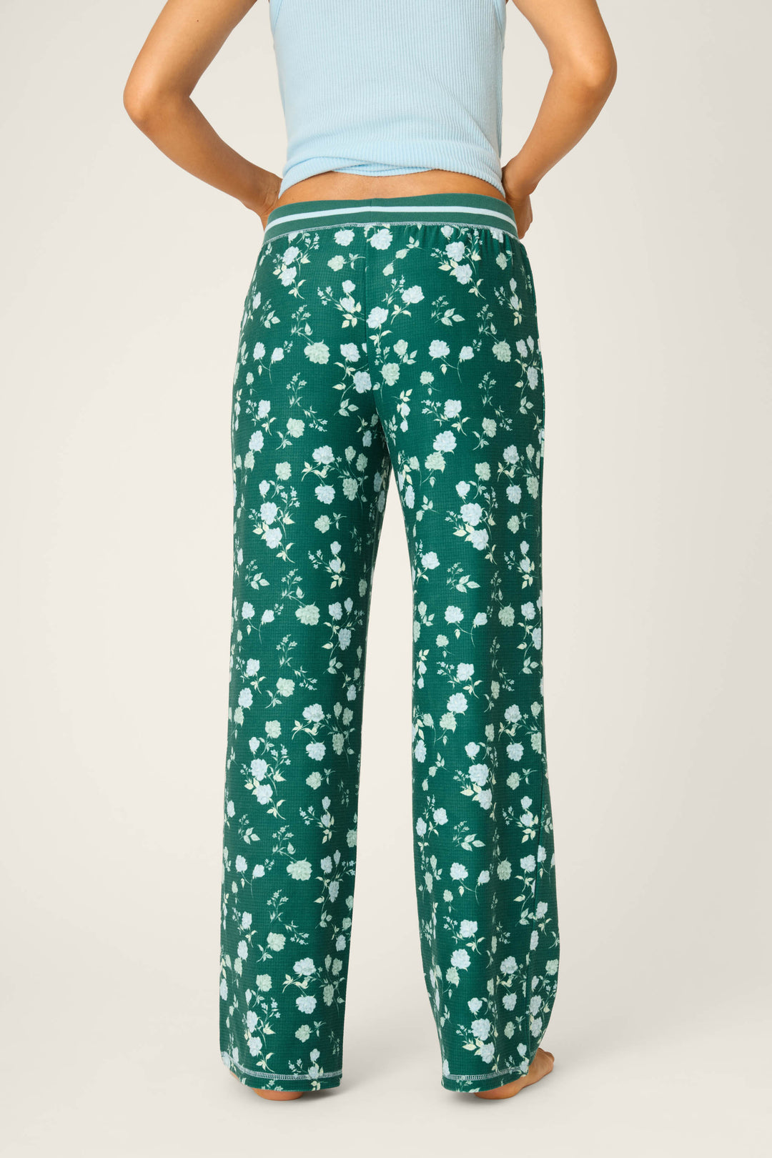 A person stands barefoot against a plain light background, seen from behind wearing P.J. Salvage Winter Wonderland Floral Thermal Velour Open Leg Pants with a white pattern and a light blue ribbed tank—ideal for cozy holiday lounging.