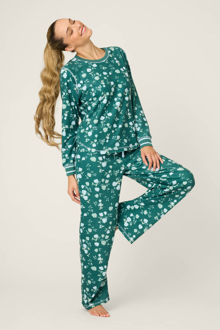 A woman with long, wavy hair smiles serenely, standing barefoot on one leg, eyes closed and head tilted back, wearing the FLORAL PJ SET WNTER LAND by P.J. Salvage—a matching green set with a white floral pattern.