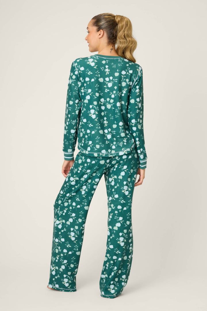 A woman faces away, wearing the P.J. Salvage FLORAL PJ SET WINTER LAND—a dark green, white floral pajama set with a long-sleeve top and loose pants. Her long hair is styled in a ponytail.