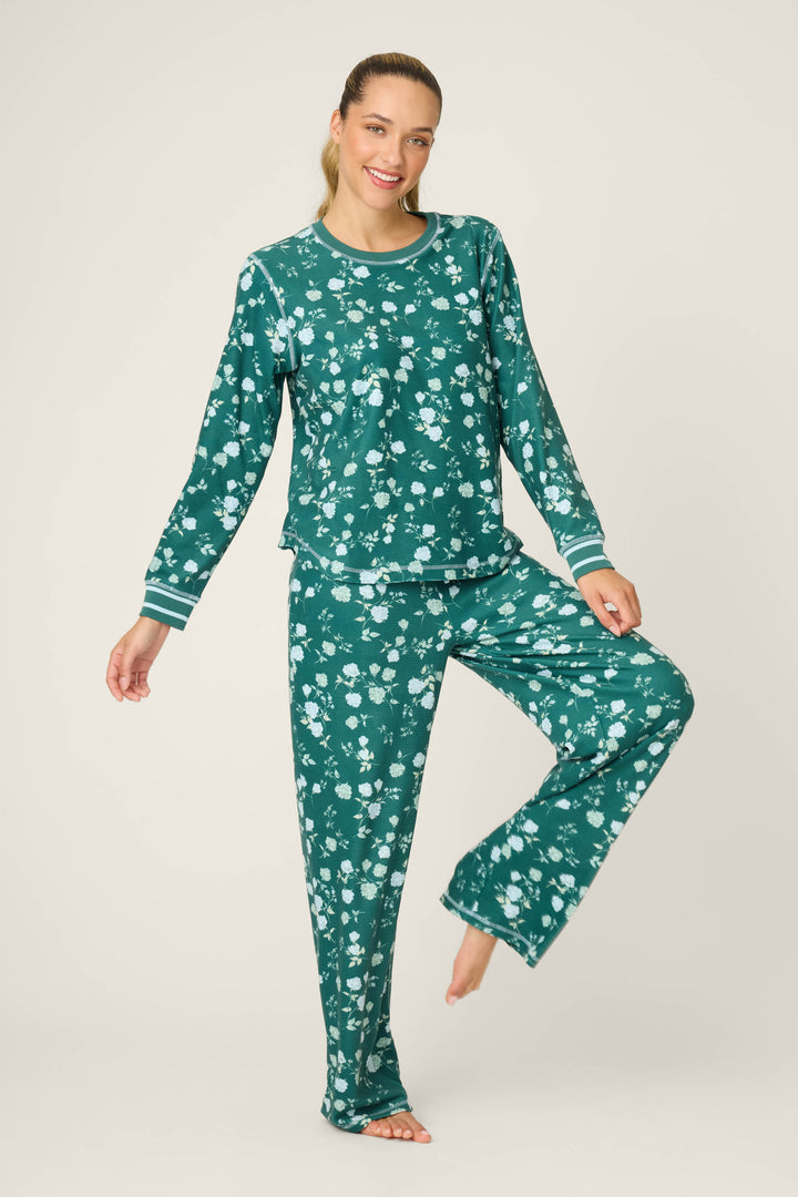A person stands on one leg, smiling, wearing the P.J. Salvage FLORAL PJ SET WINTER LAND—matching green floral print long-sleeve pajamas and pants—against a plain light background.