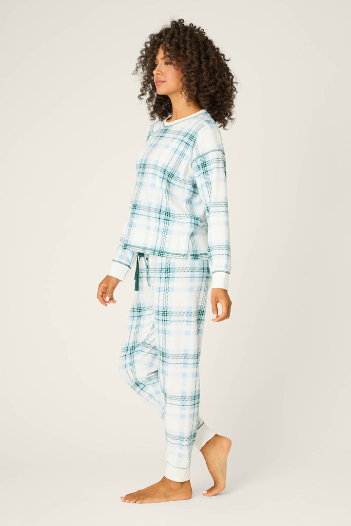 A woman with curly hair stands sideways, barefoot, modeling the P.J. Salvage Winter Wonderland Plaid Thermal Velour Pants Set in blue and white against a plain, light-colored background.