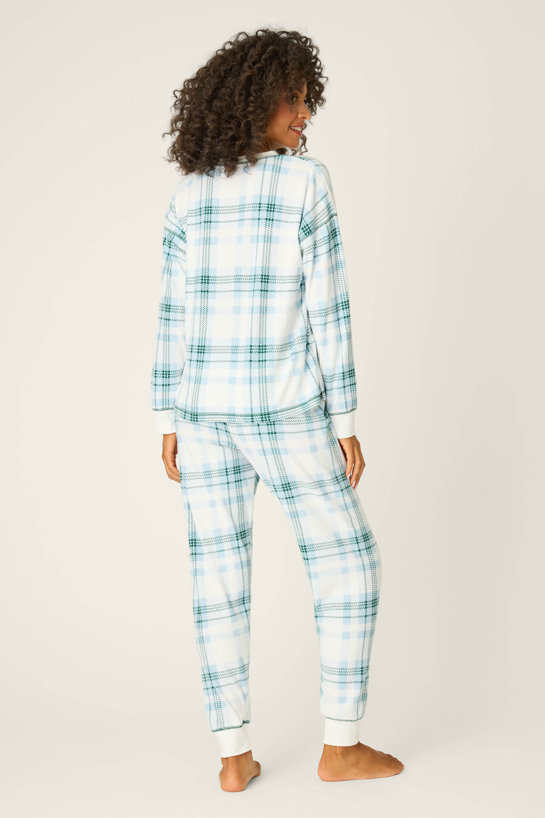 A person stands barefoot facing away, wearing the P.J. Salvage Winter Wonderland Plaid Thermal Velour Pants Set in light blue, green, and white plaid against a plain off-white background.