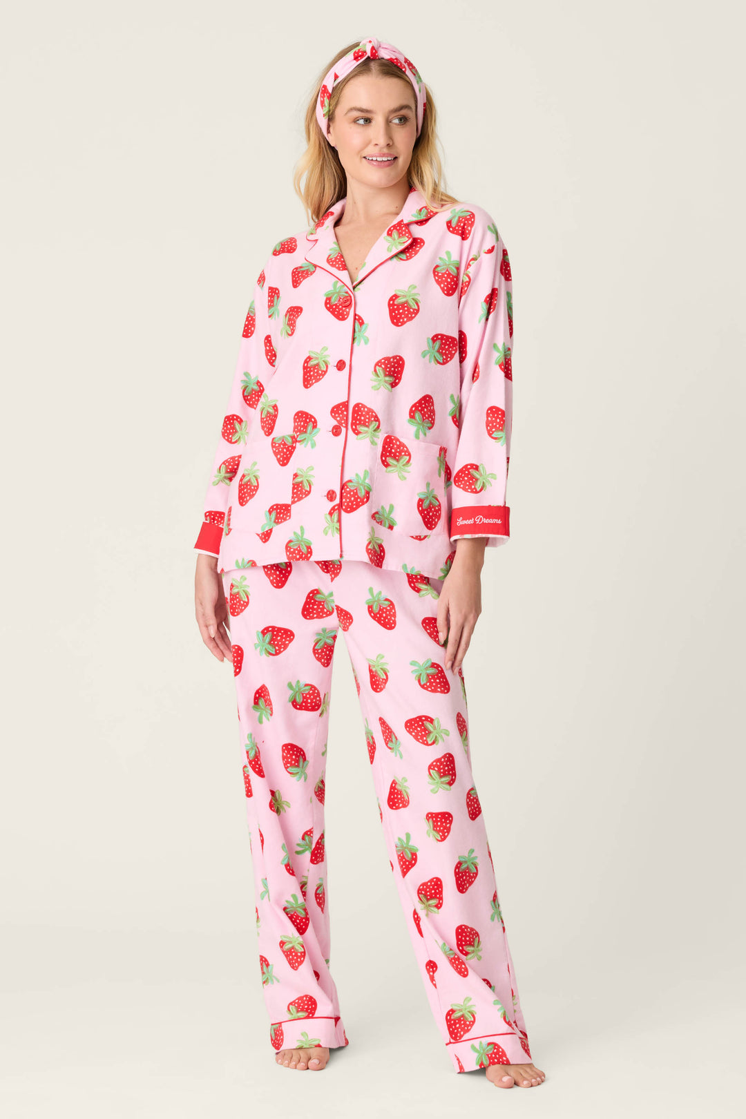 A person wears the P.J. Salvage Sweet Dreams Strawberry Print 3-Piece Flannel PJ Set, featuring pink pajamas with red strawberry print, red cuffs, a long-sleeve button-up top, matching pants, and a coordinating headband.