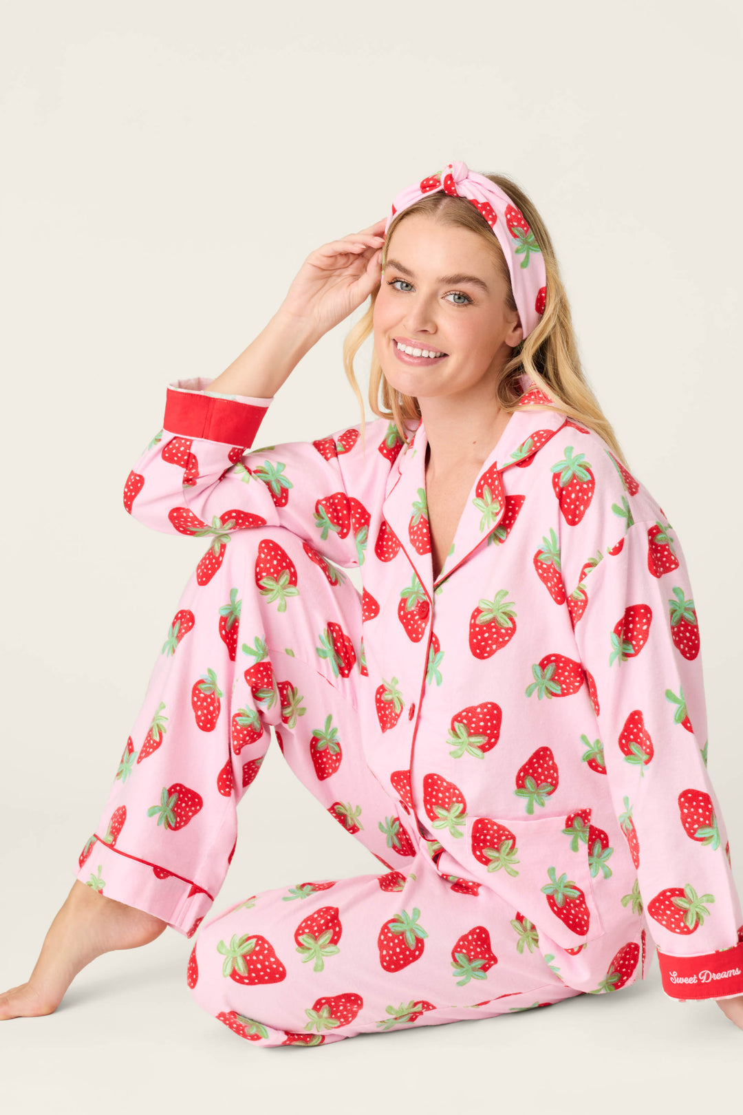 A woman sits on the floor in the P.J. Salvage Sweet Dreams Strawberry Print 3-Piece Flannel PJ Set, featuring playful pink strawberries, a matching headband, red cuffs, and “Sweet Dreams” on the wrist, smiling with one hand on her head.