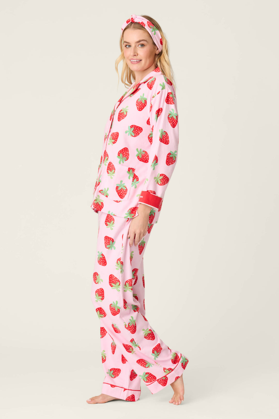 A person stands barefoot against a light background wearing the P.J. Salvage Sweet Dreams Strawberry Print 3-Piece Flannel PJ Set in pink, featuring a red strawberry print, matching headband, long-sleeved button-up top, and long pants.