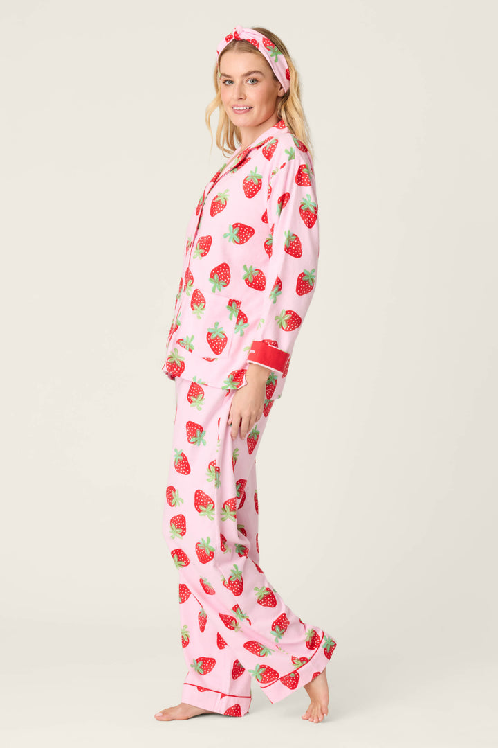 A person stands barefoot against a light background wearing the P.J. Salvage Sweet Dreams Strawberry Print 3-Piece Flannel PJ Set in pink, featuring a red strawberry print, matching headband, long-sleeved button-up top, and long pants.