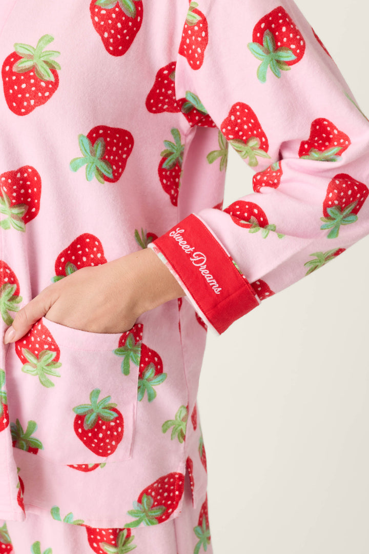 A person wears the P.J. Salvage Sweet Dreams Strawberry Print 3-Piece Flannel PJ Set with pink fabric, strawberry print, a red inner cuff on the sleeve reading “Sweet Dreams” in white script, and their left hand tucked into a pocket.
