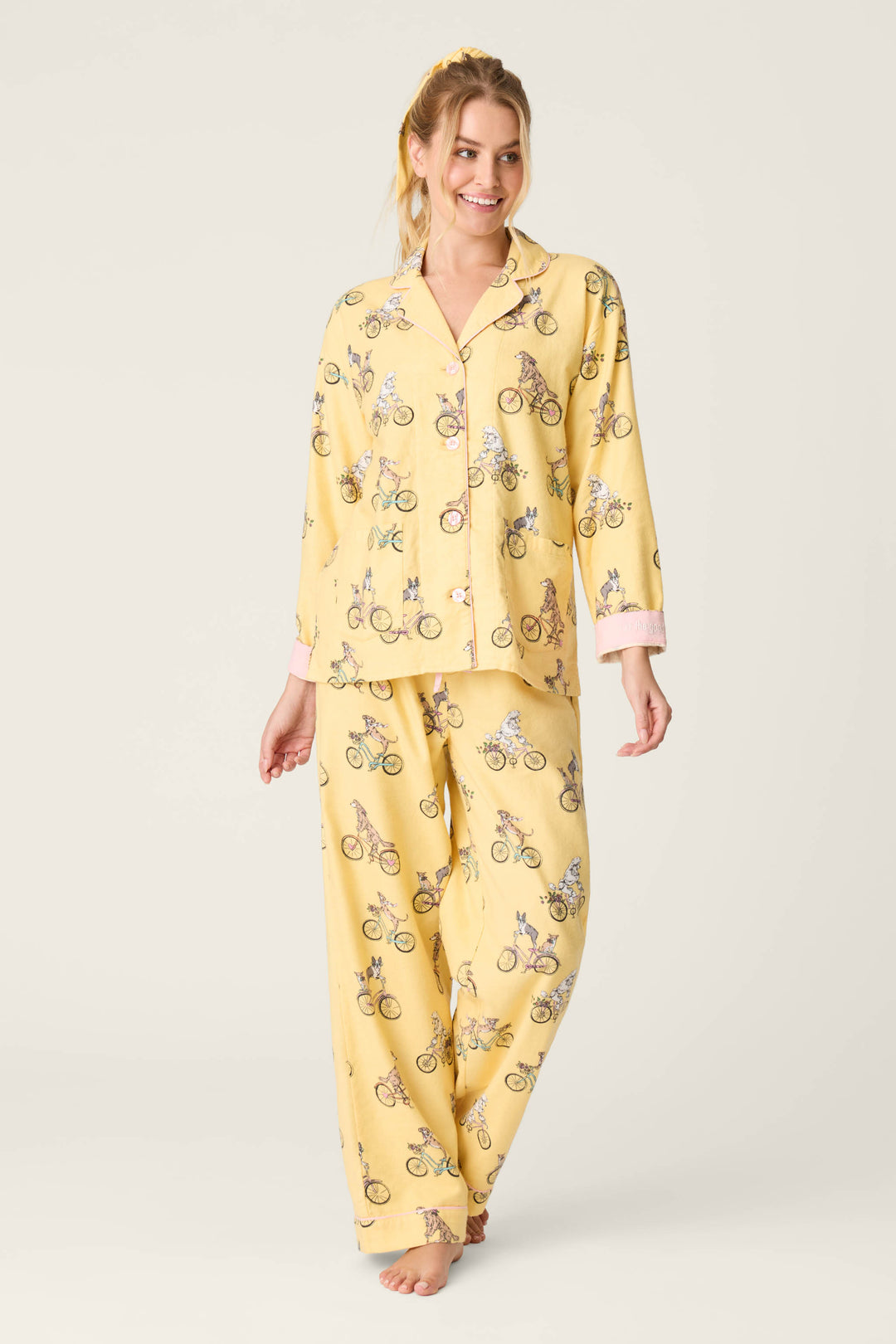 A woman, barefoot and smiling, wears the P.J. Salvage "Let the Good Times Roll" 3-piece flannel PJ set: a yellow long-sleeve buttoned top and loose bicycle-print pants. She looks to the side against a plain background.