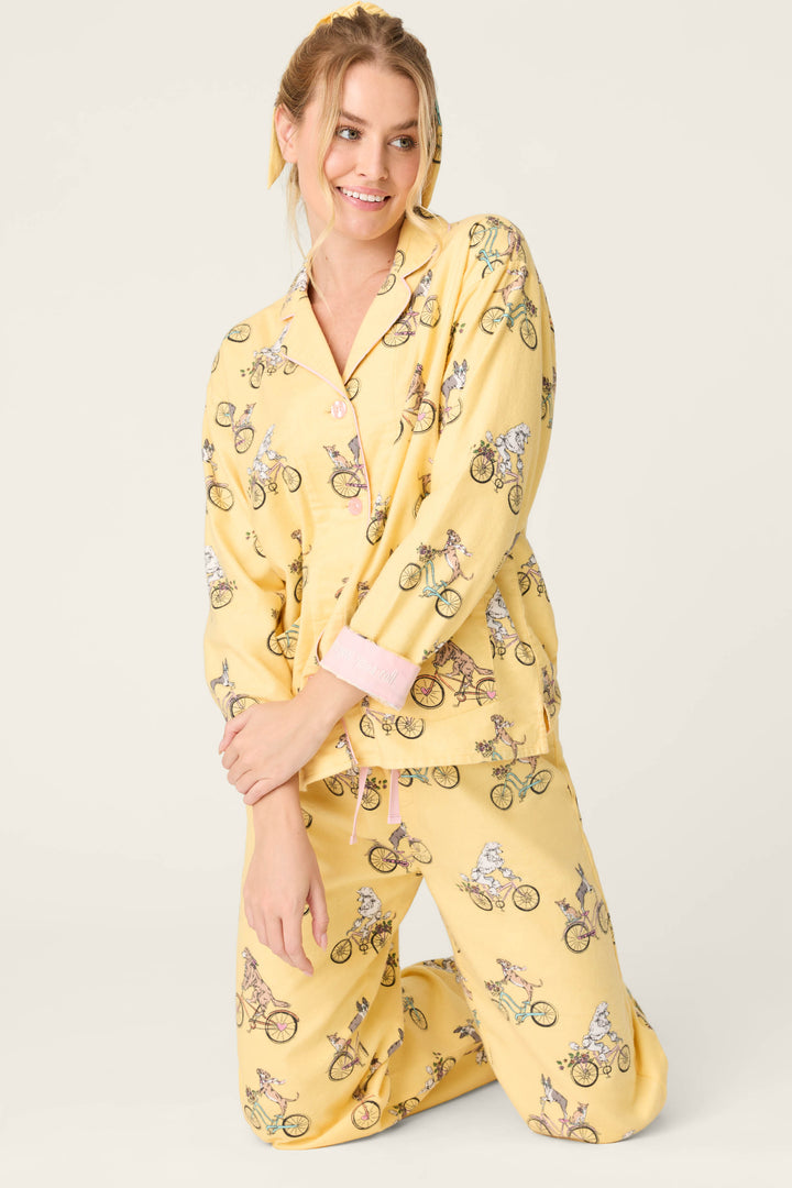 A person kneels on one knee, smiling in the P.J. Salvage Let the Good Times Roll 3-Piece Flannel PJ Set—yellow with dogs riding bicycles and pink piping, exuding cozy sleepwear vibes against a plain, light-colored background.