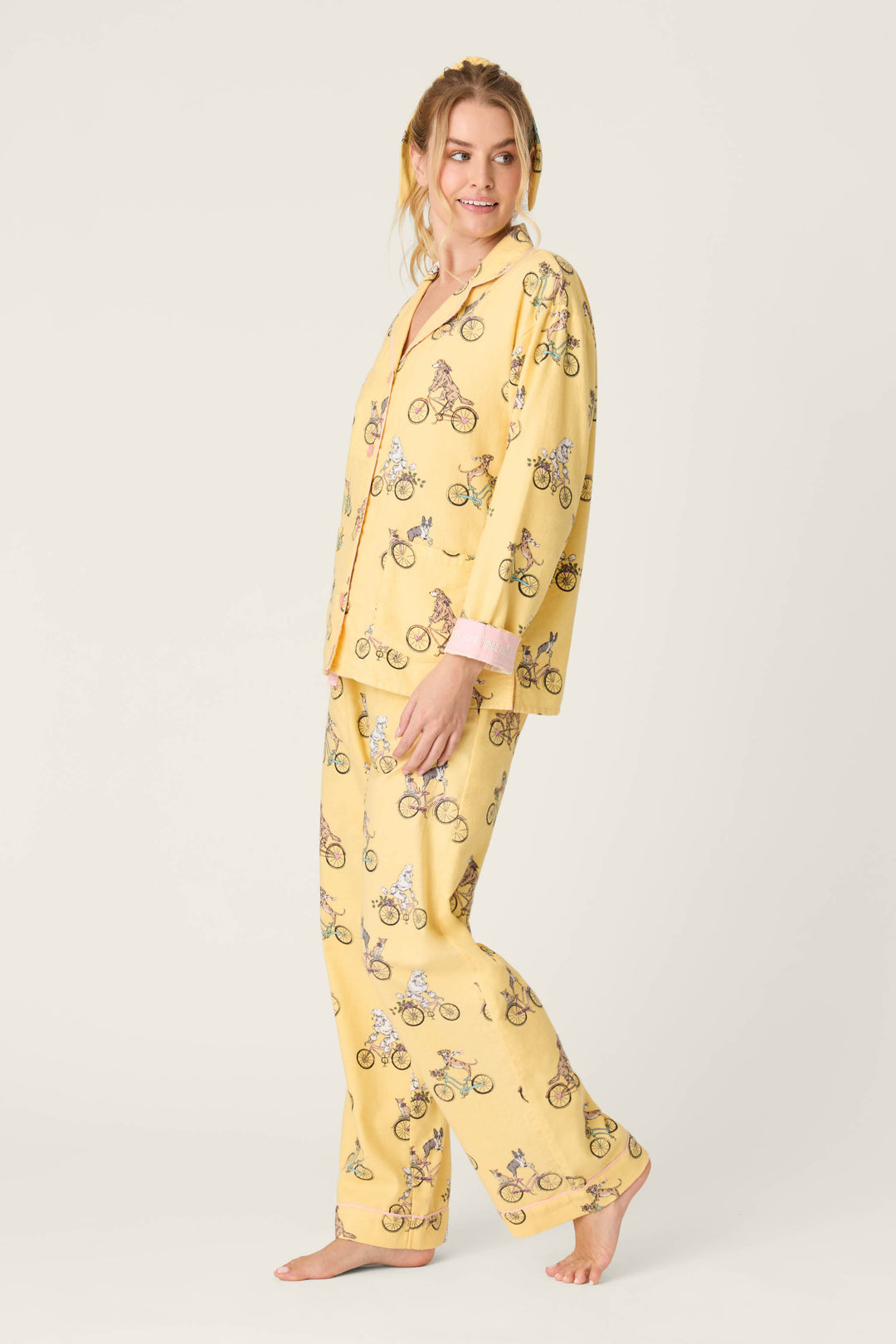 A woman stands barefoot against a plain background, smiling as she models the P.J. Salvage Let the Good Times Roll 3-Piece Flannel PJ Set—yellow sleepwear with a bicycle print, including a long-sleeved button-up top and loose-fit pants.