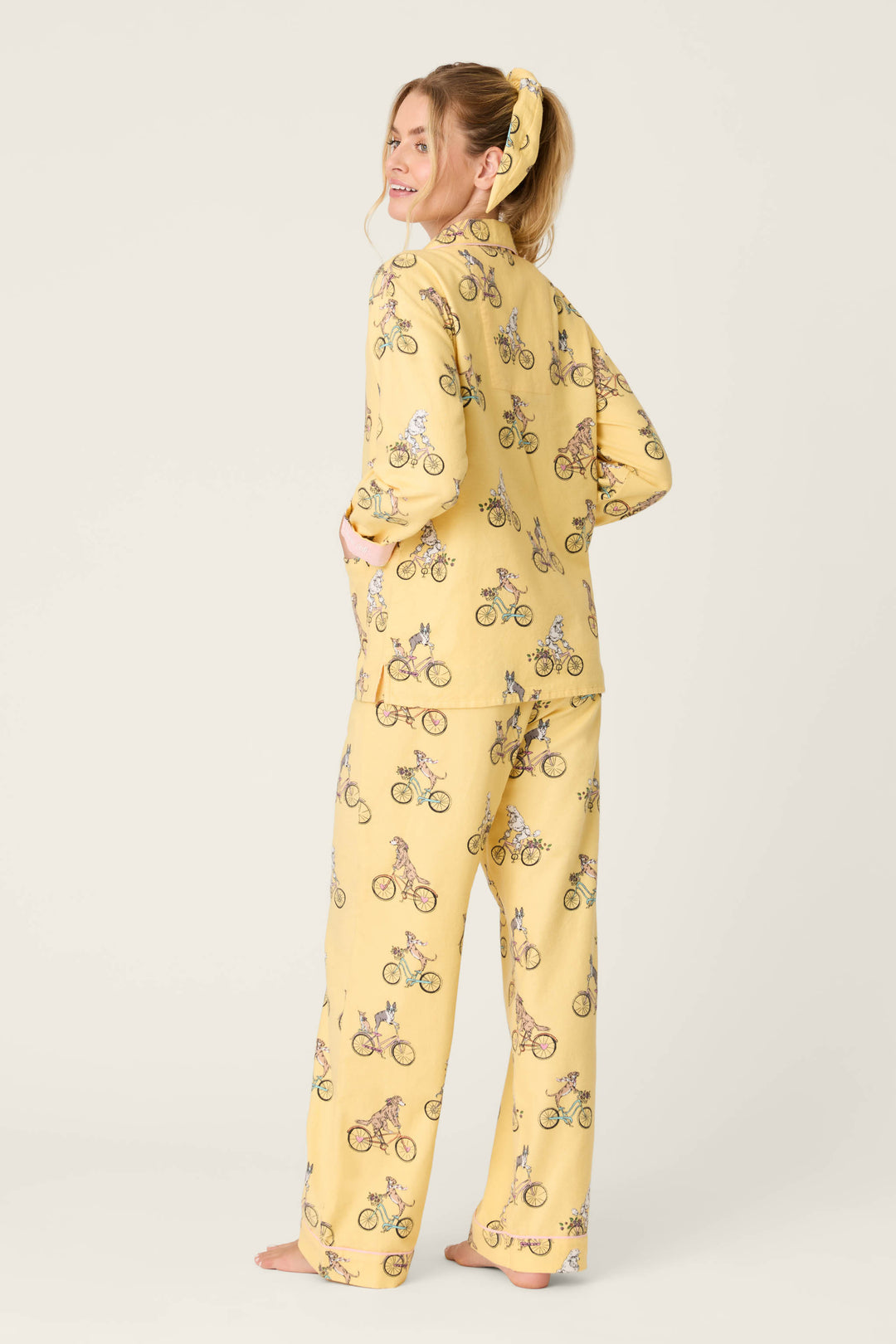 A person stands barefoot on a light background, facing away and smiling over their shoulder, wearing the P.J. Salvage "Let the Good Times Roll" 3-Piece Flannel PJ Set in yellow with a bicycle print and matching scrunchie.