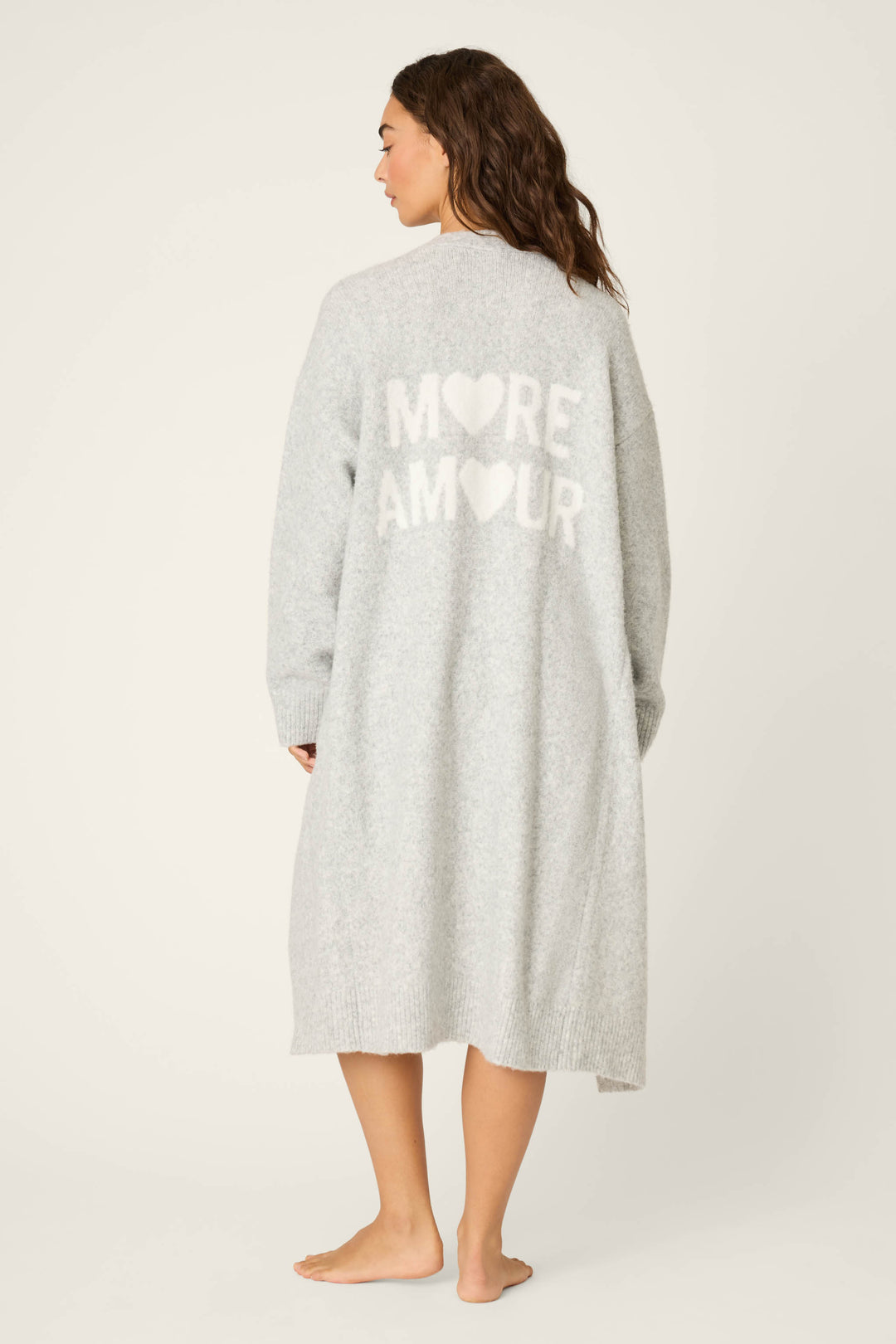 A person stands barefoot on a light background, facing away to show the cozy texture and relaxed fit of the P.J. Salvage My Dog Is My Valentine Long Line Cardigan in heather grey, featuring white heart designs and MORE AMOUR text on the back.