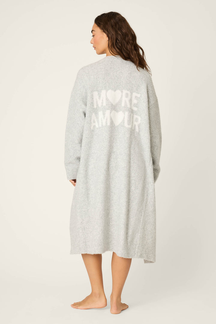 A person stands barefoot on a light background, facing away to show the cozy texture and relaxed fit of the P.J. Salvage My Dog Is My Valentine Long Line Cardigan in heather grey, featuring white heart designs and MORE AMOUR text on the back.