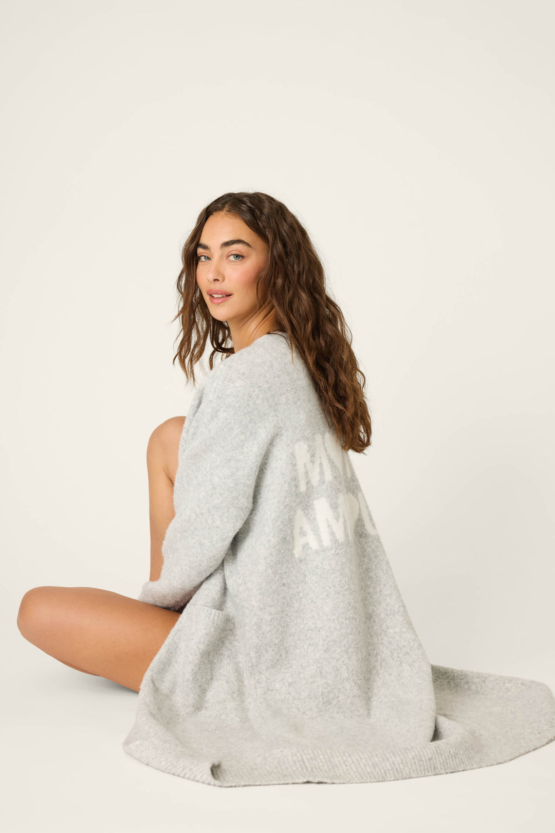 A woman with long, wavy brown hair sits against a plain background wearing the P.J. Salvage My Dog Is My Valentine Long Line Cardigan in heather grey. The cozy, oversized cardigan drapes over her legs and features partially visible white text on the back.