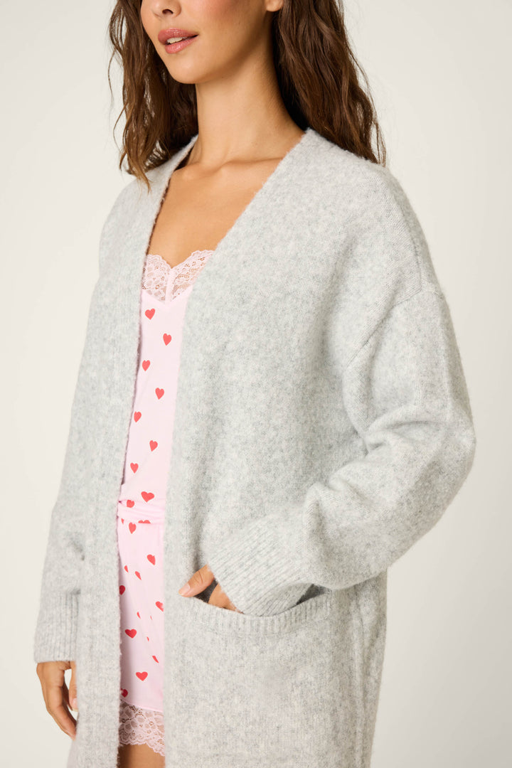 A woman wears the P.J. Salvage "My Dog Is My Valentine" Long Line Cardigan in heather grey over a light pink camisole and red heart-print shorts, standing with one hand in her cardigan pocket, facing slightly to the side.