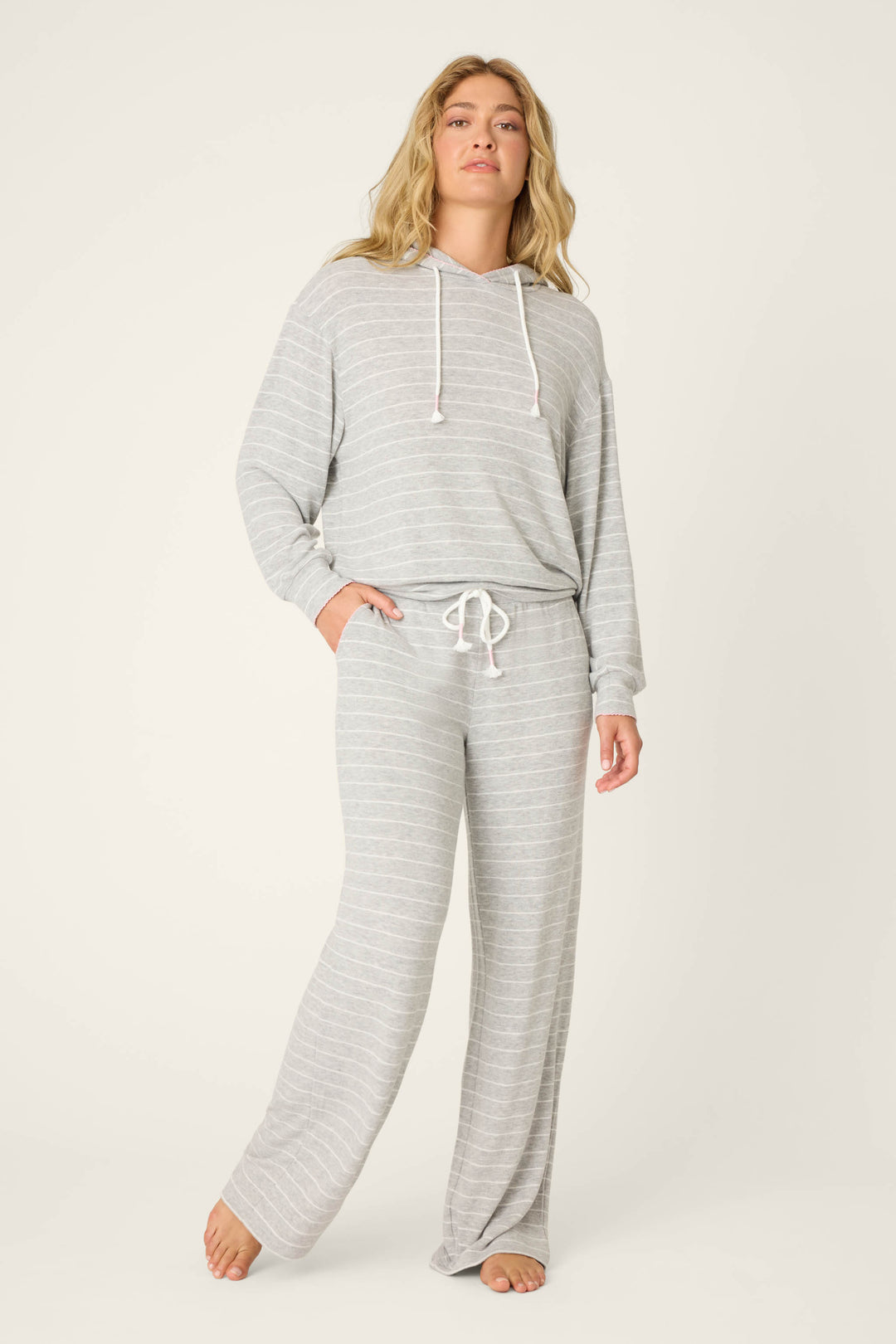 A person stands barefoot against a plain background, wearing the P.J. Salvage My Dog Is My Valentine Striped Hoodie lounge set in light grey with white drawstrings and thin stripes, their left hand tucked into a pocket.