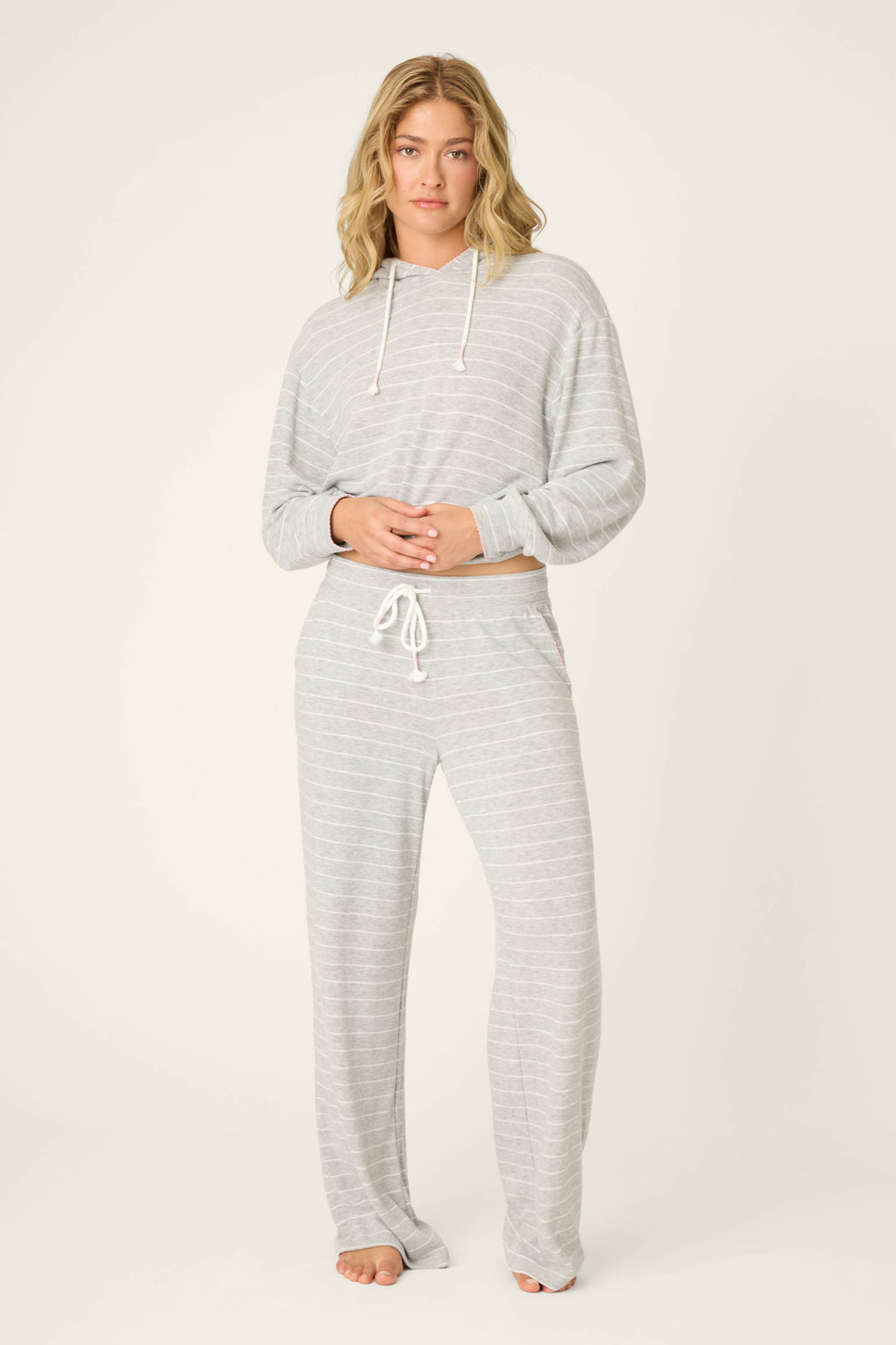 A person stands barefoot against a plain background, wearing P.J. Salvage’s My Dog Is My Valentine Striped Open Leg Pants—a light gray, drawstring style with subtle white stripes—paired with a matching hooded top and their hands clasped in front.