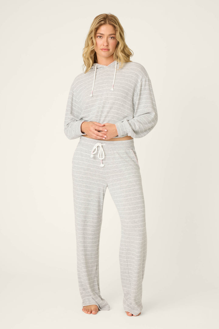 A person stands barefoot against a plain background, wearing P.J. Salvage’s My Dog Is My Valentine Striped Open Leg Pants—a light gray, drawstring style with subtle white stripes—paired with a matching hooded top and their hands clasped in front.