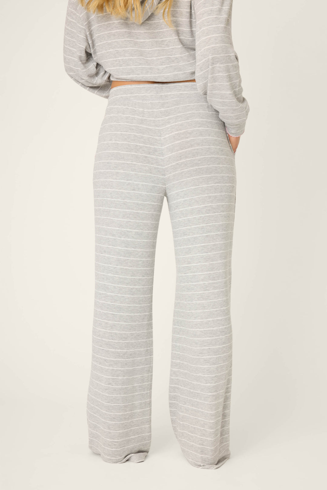A woman faces away from the camera, wearing P.J. Salvage’s My Dog Is My Valentine Striped Open Leg Pants in light gray with subtle stripes and a high waist, paired with a matching long-sleeve top. Her right hand is in her pocket.