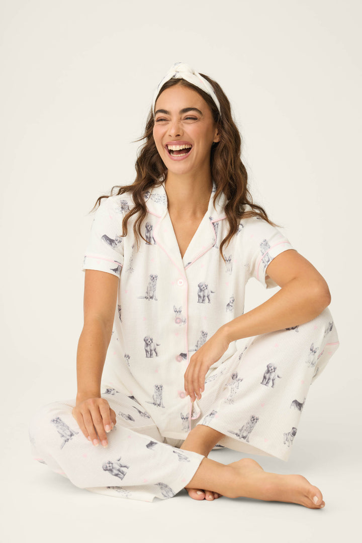 A woman in the P.J. Salvage "My Dog Is My Valentine" 3-piece PJ set, featuring a dog print, sits cross-legged on the floor with a white headband and smiles off to the side against a plain light background.