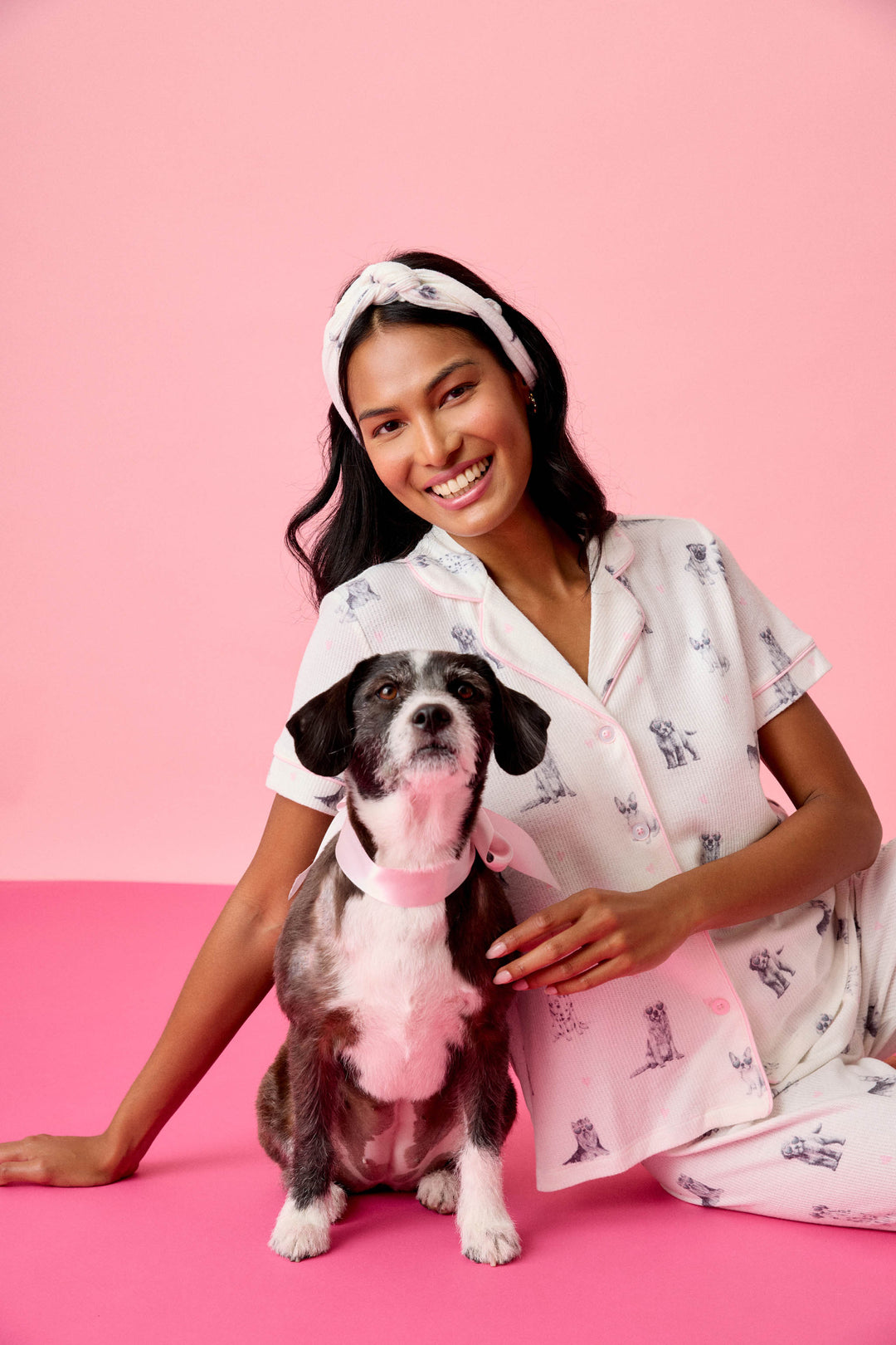 A woman in the P.J. Salvage "My Dog Is My Valentine" 3-Piece PJ Set sits on a pink floor, smiling at the camera with one arm around a black and white dog wearing a light pink collar, against a pink background.