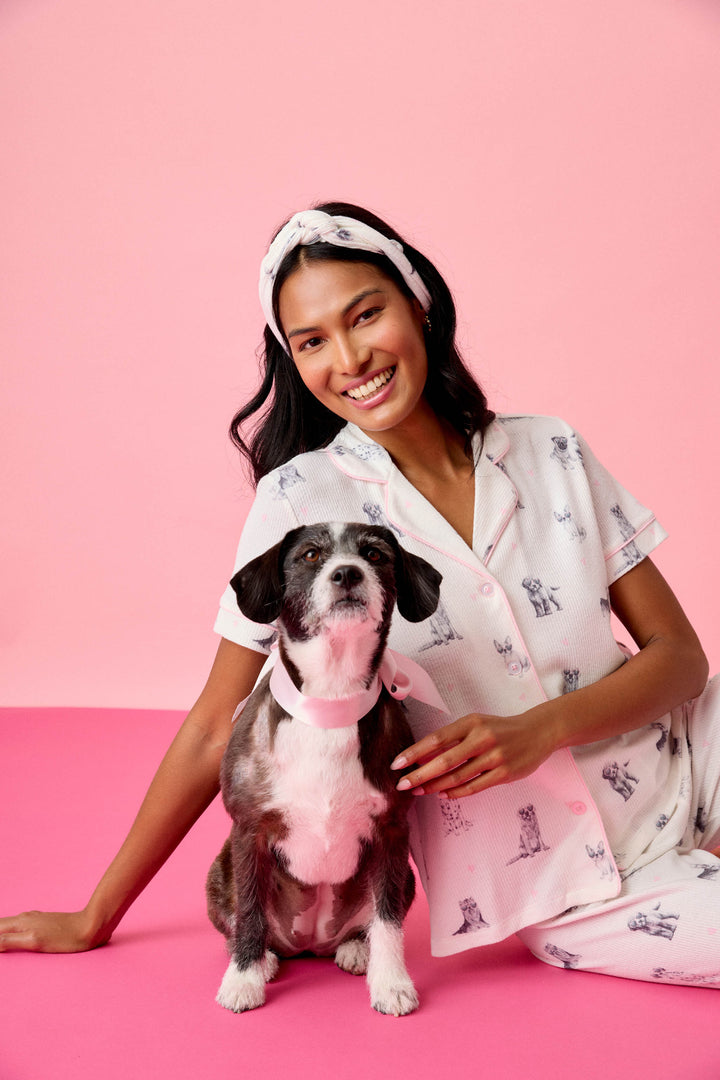 A woman in the P.J. Salvage "My Dog Is My Valentine" 3-Piece PJ Set sits on a pink floor, smiling at the camera with one arm around a black and white dog wearing a light pink collar, against a pink background.