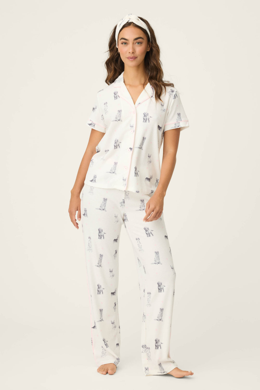 A woman with long hair stands barefoot in the P.J. Salvage "My Dog Is My Valentine" 3-piece PJ set—featuring a white, dog-printed short-sleeve button-up top, long pants, and matching headband—against a plain, light background.