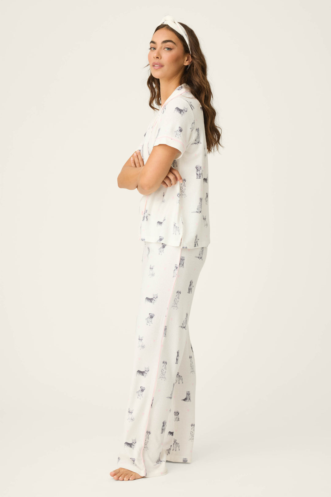 A woman with long brown hair models the P.J. Salvage "My Dog Is My Valentine" 3-piece PJ set—white with animal prints and a matching headband—standing barefoot, arms crossed, facing slightly to the side against a plain light background.
