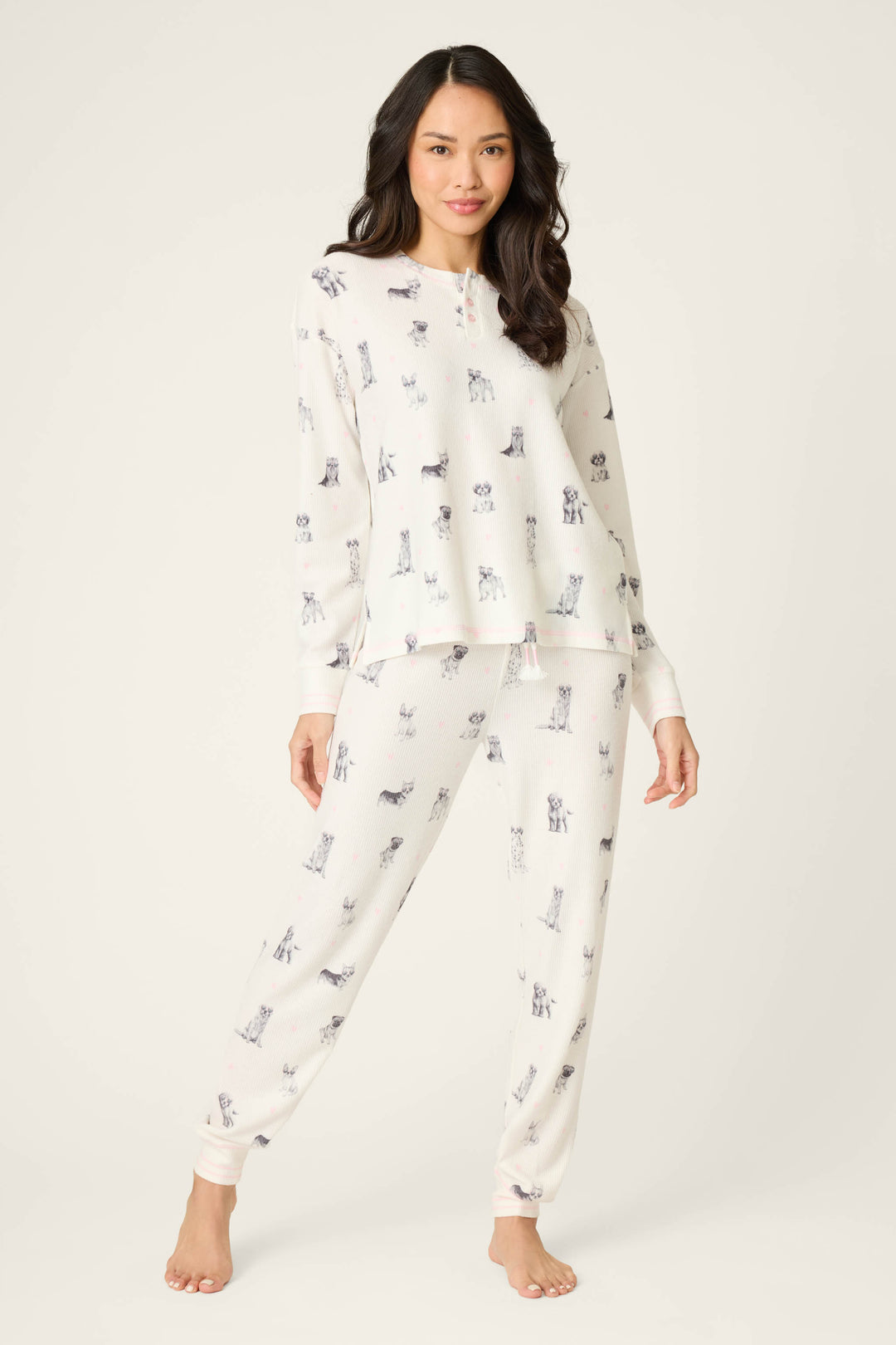 A woman with long dark hair stands barefoot on a light background, wearing the P.J. Salvage "My Dog Is My Valentine" PJ set, which features a matching white pajama design with a dog print. She faces forward and smiles slightly.