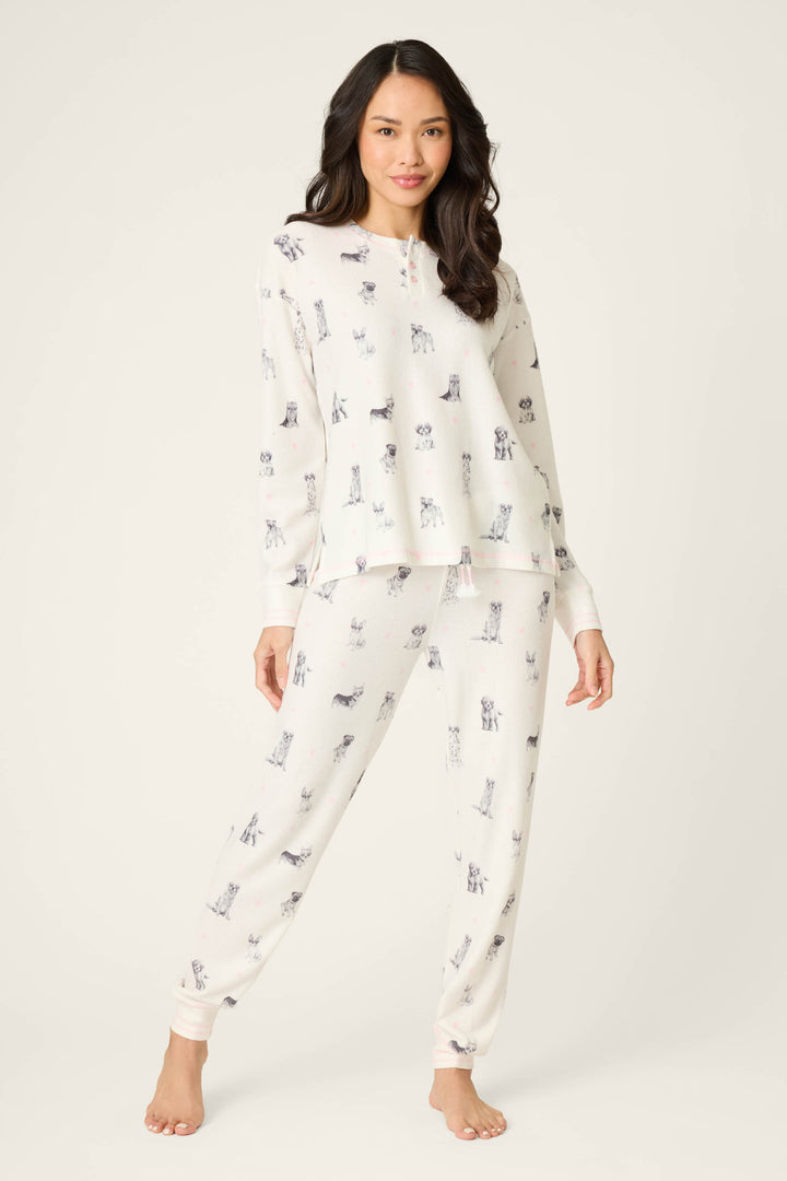 A woman with long dark hair stands barefoot on a light background, wearing the P.J. Salvage "My Dog Is My Valentine" PJ set, which features a matching white pajama design with a dog print. She faces forward and smiles slightly.