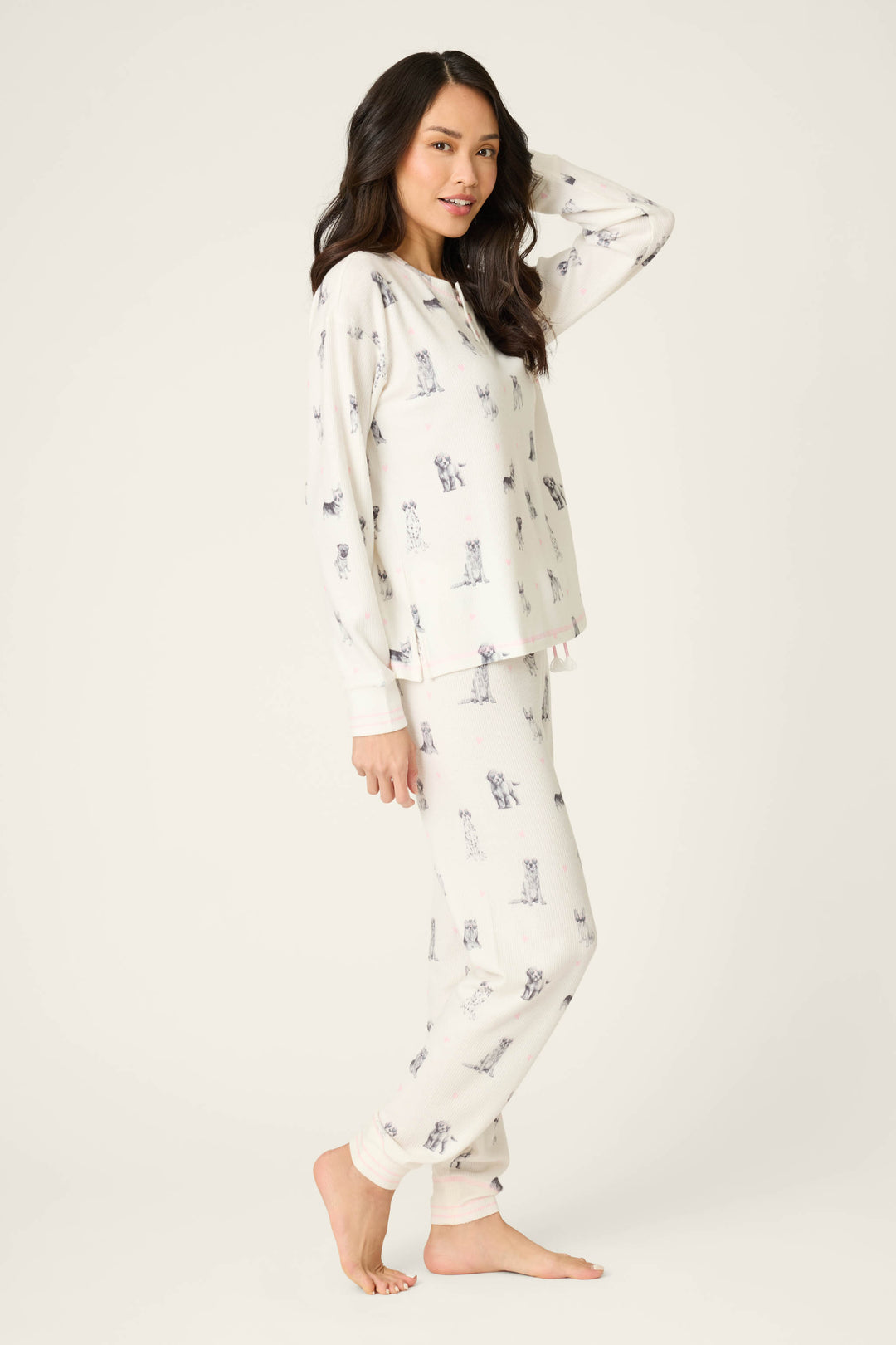 A woman stands barefoot on a plain background, wearing the P.J. Salvage My Dog Is My Valentine PJ Set featuring a dog print. She has long dark hair, faces slightly sideways, and looks at the camera with a relaxed expression.