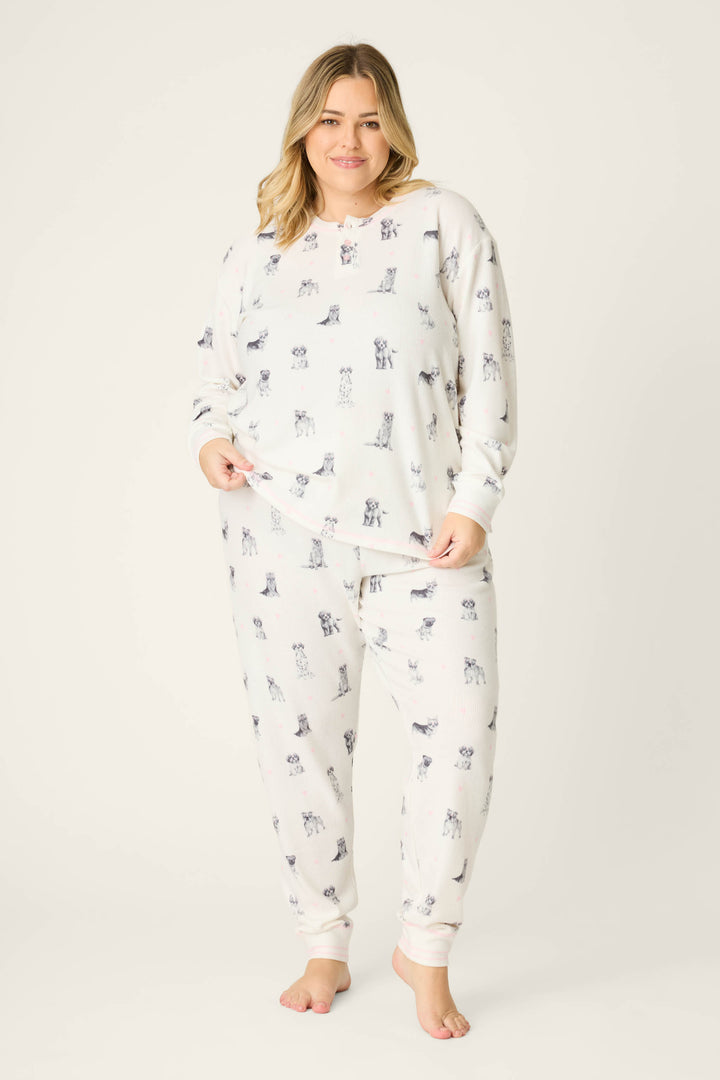 A woman stands barefoot on a plain background, smiling slightly as she models the P.J. Salvage My Dog Is My Valentine Plus Size PJ Set, featuring a dog print pattern, her hands resting on the top and side of her pants.
